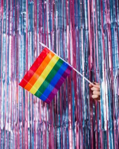 This is a picture of a pride flag held infront of a tinsel background which are pale pink and blue, the colours of the trans flag. 